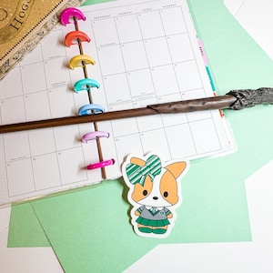 May include: A planner with colorful dividers and a wand. A sticker of a cartoon corgi wearing a green and white bow and a school uniform is on the bottom. The planner is open to a calendar page.