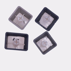 K&ouml;nnte beinhalten: Vier Tastaturkappen. Zwei sind dunkelgrau und zwei hellgrau. Jede Tastenkappe hat einen quadratischen Rahmen mit einer kreuzf&ouml;rmigen Halterung im Inneren. Die Tastenkappen werden von oben vor einem wei&szlig;en Hintergrund betrachtet.