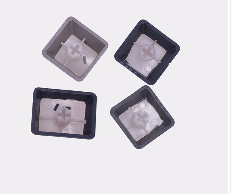 K&ouml;nnte beinhalten: Vier Tastaturkappen. Zwei sind dunkelgrau und zwei hellgrau. Jede Tastenkappe hat einen quadratischen Rahmen mit einer kreuzf&ouml;rmigen Halterung im Inneren. Die Tastenkappen werden von oben vor einem wei&szlig;en Hintergrund betrachtet.