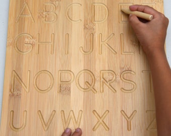 May include: A wooden alphabet tracing board with a child's hand tracing the letters with a wooden stylus. The board has the entire alphabet in uppercase letters.
