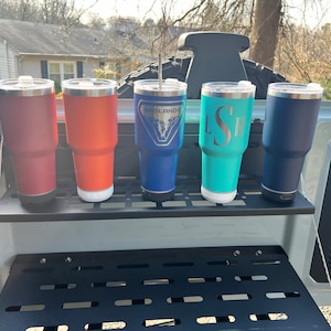 May include: Five stainless steel tumblers with lids and various colors, including red, orange, blue, teal, and navy blue. The tumblers are arranged on a black metal shelf with a grid pattern. The tumblers have different designs, including a logo that says "Badlands" and a monogram that says "LSH".