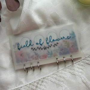 May include: A white, translucent, six-ring binder with a floral design and the text "field of flowers" in teal lettering.