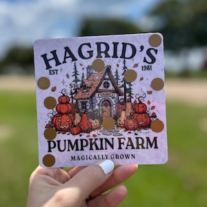May include: A square, light purple sign with the words "HAGRID'S PUMPKIN FARM" and a graphic of a cottage and pumpkins. The sign also has gold circles and the text "MAGICALY GROWN".