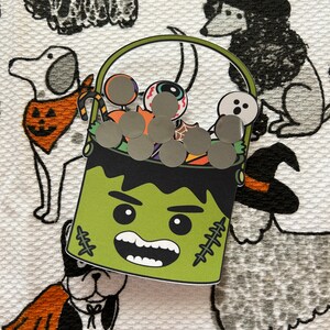 May include: A green Frankenstein-themed Halloween candy bucket with a black handle, filled with gray circular candies and Halloween-themed candies. The bucket is on a white patterned surface with dog illustrations.