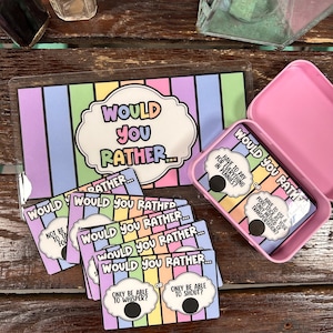 May include: A Would You Rather game set with cards and a pink tin. The cards feature colorful rainbow stripes and the text "Would You Rather..." with various choices. The tin is open, revealing more cards with similar designs.