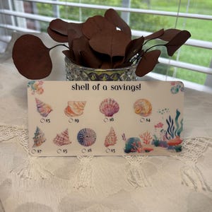 May include: A decorative card with the text "shell of a savings!" features colorful seashell illustrations. The card includes various seashell designs with prices listed below each. The card is on a white lace tablecloth.
