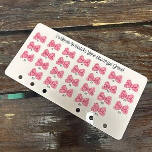 May include: A white savings tracker card with 26 pink bow illustrations, each with a dollar amount. The text "26 Bows to Watch Your Savings Grow!" is printed at the top. The card has three holes for binder use.