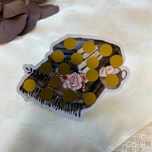 May include: A sticker featuring a cozy scene with books, a blanket, flowers, and a mug of hot chocolate. Gold foil circles are scattered across the image. The sticker has a white border and is set against a white background.