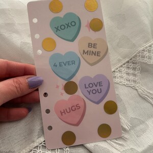 May include: A pink planner insert with heart-shaped candies that say "XOXO", "BE MINE", "4 EVER", "LOVE YOU", and "HUGS". Gold circle stickers and pink star accents are also on the insert.