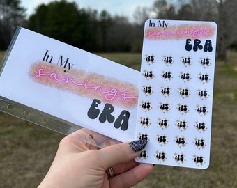 May include: A white sticker sheet with the text "In My Savings Era" in pink and black lettering. The sticker sheet has 25 stickers with the number "$1.5" and a gold star on each sticker.