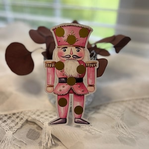 May include: A pink and gold nutcracker sticker with a white beard and mustache. The sticker has a pink hat with gold dots and a pink uniform with gold accents. The sticker is placed on a white surface with a lace trim.