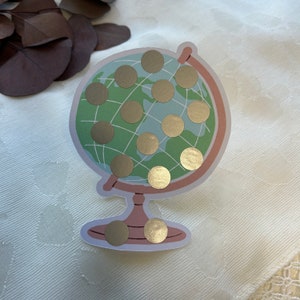 May include: A sticker in the shape of a globe with a light green background and gold foil circles. The globe has a light brown frame and base. The sticker is on a white surface with a subtle pattern.
