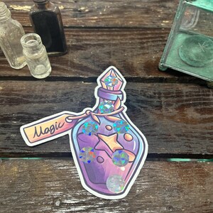 May include: A sticker of a purple potion bottle with a star and holographic gems. The bottle has a label that says "Magic". Other small glass bottles are in the background.