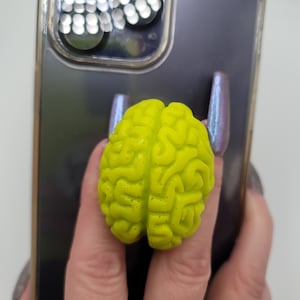 May include: A neon yellow brain-shaped phone grip is attached to a black phone case. The brain grip has detailed ridges and a textured surface. The phone case has a clear border with decorative elements.