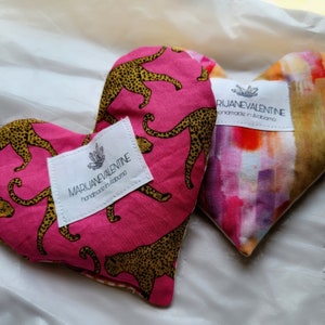May include: Two heart-shaped fabric heat packs with leopard print and abstract watercolor designs. Both packs have a white label with the text "MARIJANEVALENTINE handmade in Alabama".