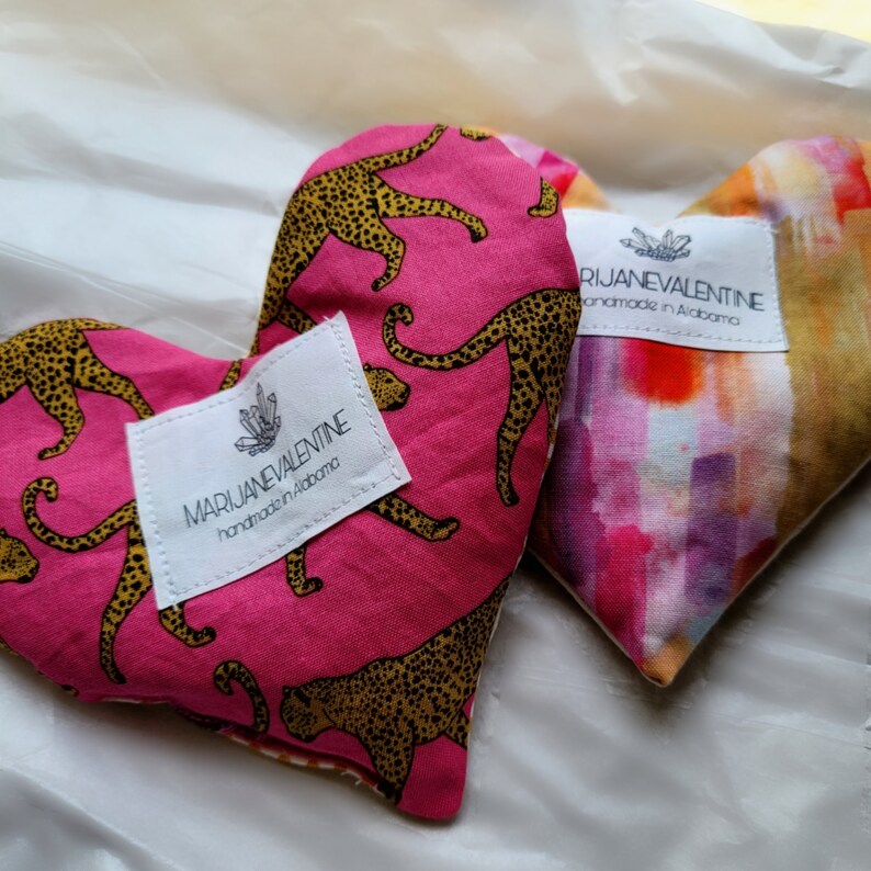 May include: Two heart-shaped fabric heat packs with leopard print and abstract watercolor designs. Both packs have a white label with the text "MARIJANEVALENTINE handmade in Alabama".