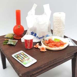 May include: A messy table with a red glass bong, a plastic bag, a roll of paper towels, a plate of food, a can of Coca-Cola, a cell phone, and a few other items.