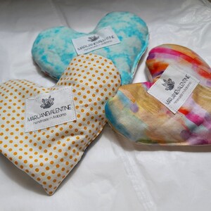 May include: Three heart-shaped pillows with different patterns. The first pillow is yellow with white polka dots, the second is blue with a white tie-dye pattern, and the third is a rainbow watercolor pattern. Each pillow has a white label with the text "MARJANEVALENTINE handmade in Alabama".