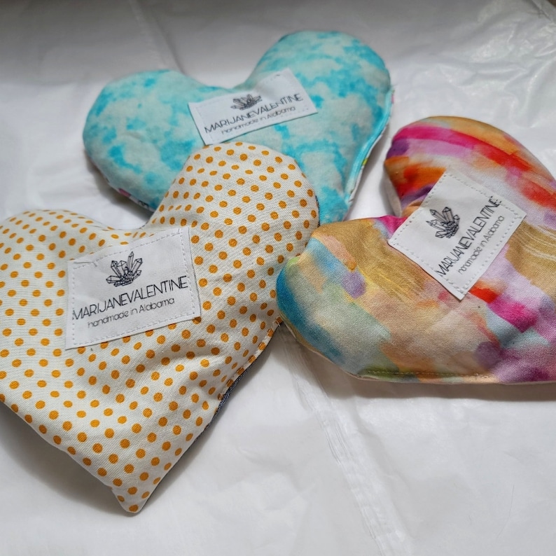 May include: Three heart-shaped pillows with different patterns. The first pillow is yellow with white polka dots, the second is blue with a white tie-dye pattern, and the third is a rainbow watercolor pattern. Each pillow has a white label with the text "MARJANEVALENTINE handmade in Alabama".