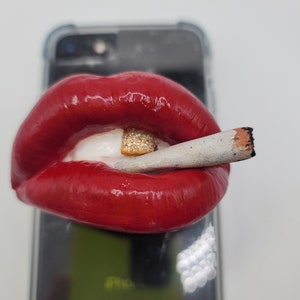 May include: A red, glossy, sculpted lip phone stand with a white tooth and a gold, diamond-encrusted tooth holding a lit cigarette.
