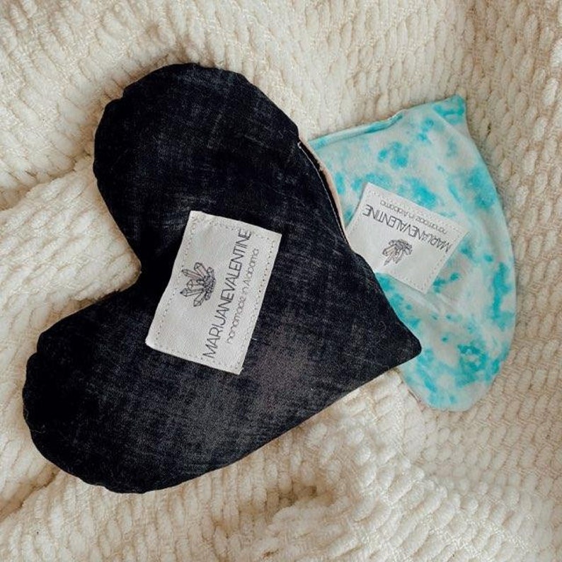 May include: Two heart-shaped fabric pillows with tie-dye patterns. The black pillow has a label that reads "MARIJUANEVALENTINE Handmade in Alabama".