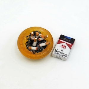 May include: A miniature yellow ashtray filled with cigarette butts and a pack of Marlboro cigarettes.