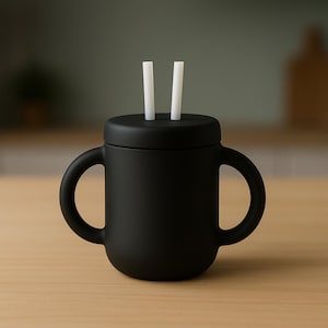 May include: A black sippy cup with two handles and a lid featuring two white straws. The cup is made of a matte material and sits on a light-coloured wooden surface. The background is a muted green.