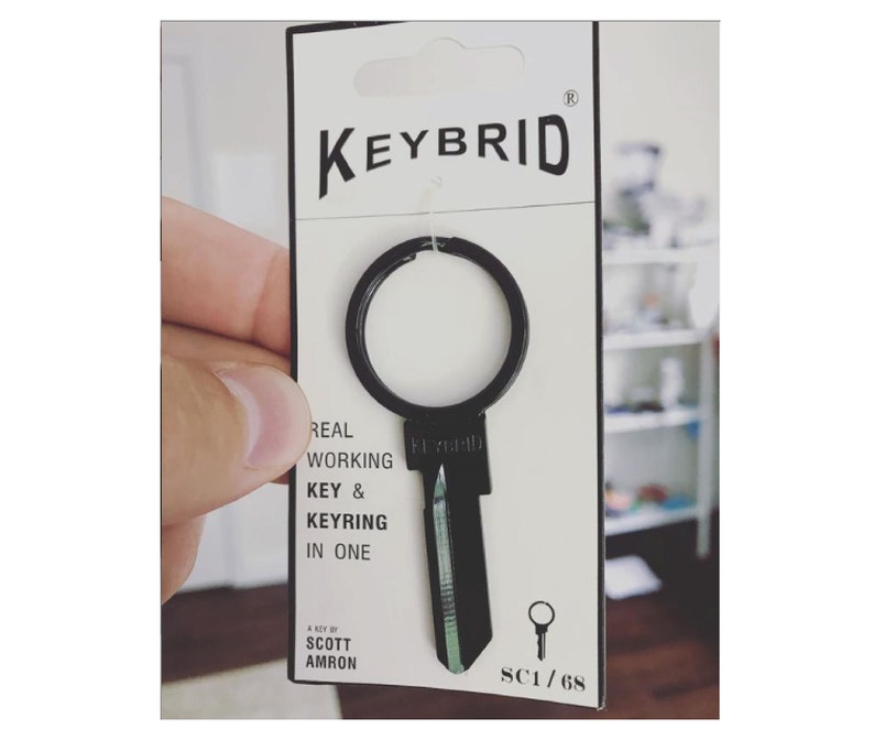 K&ouml;nnte beinhalten: Ein schwarzer schl&uuml;sself&ouml;rmiger Schl&uuml;sselanh&auml;nger mit einem Schl&uuml;sselring. Der Schl&uuml;sselanh&auml;nger tr&auml;gt den Text "KEYBRID". Die Verpackung lautet "REAL WORKING KEY & KEYRING IN ONE" und "A KEY BY SCOTT AMRON". Die Verpackung tr&auml;gt auch den Text "SC1/68".