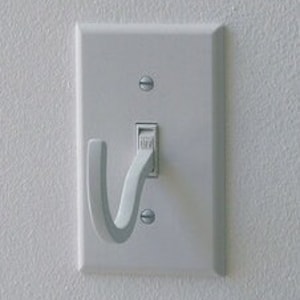 Könnte beinhalten: Ein weißer Lichtschalter mit einem weißen Plastikhaken, der an der Unterseite befestigt ist. Der Haken ist U-förmig.