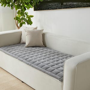 May include: A white couch with a gray quilted cushion on top. The couch has a white fabric covering and a simple design.