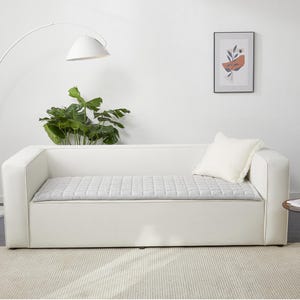 May include: A white sofa with a gray quilted cushion and a white throw pillow. The sofa is in a room with a white floor and a white wall. There is a plant in a pot on the floor to the left of the sofa.