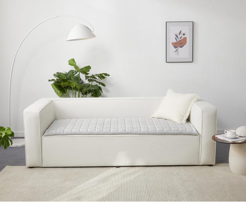 May include: A white sofa with a gray quilted cushion and a white throw pillow. The sofa is in a room with a white floor and a white wall. There is a plant in a pot on the floor to the left of the sofa.