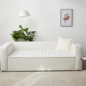 May include: White sofa with a quilted cushion and a white throw pillow. The sofa is in a white room with a floor lamp and a framed print on the wall.