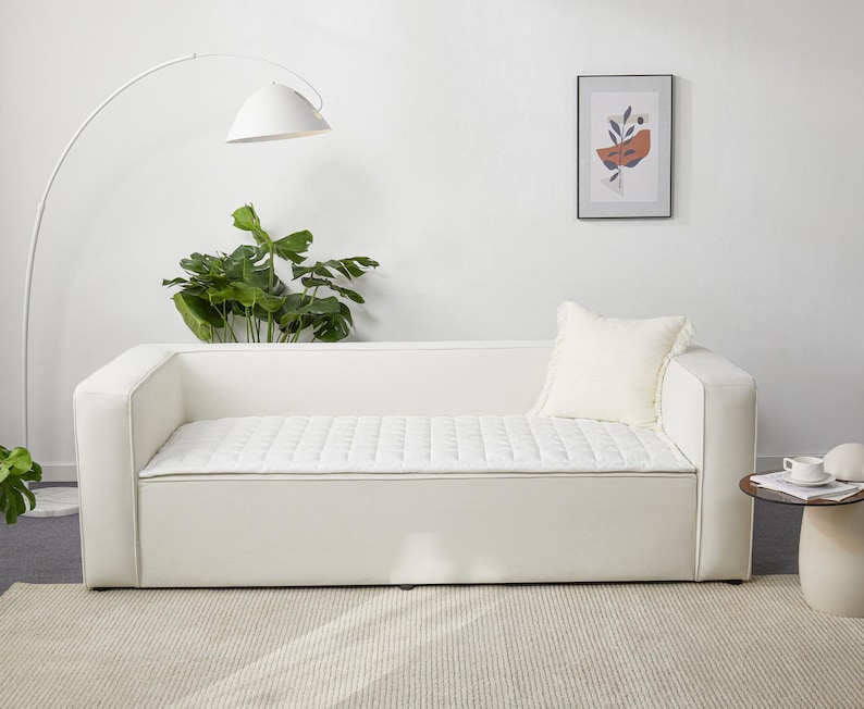 May include: White sofa with a quilted cushion and a white throw pillow. The sofa is in a white room with a floor lamp and a framed print on the wall.