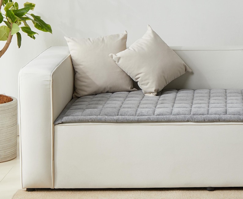May include: A white couch with a gray quilted cushion. Two white pillows are on the couch.