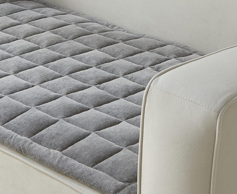 May include: Gray quilted sofa protector with a diamond pattern. The protector is placed on a white sofa.