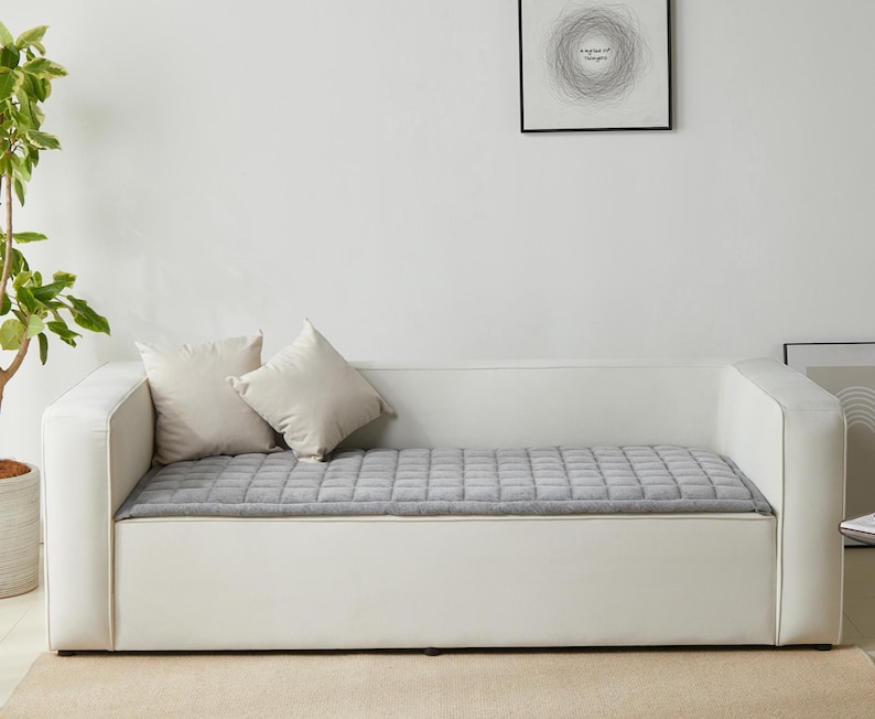 May include: A white sofa bed with a gray quilted mattress and two white pillows. The sofa bed is against a white wall with a framed print above it.