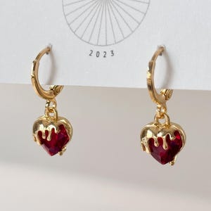May include: A pair of gold hoop earrings featuring dangling heart-shaped charms. Each charm consists of a deep red faceted heart encased in a gold-toned metal frame that mimics a dripping liquid effect. The earrings are attached to a white display card.