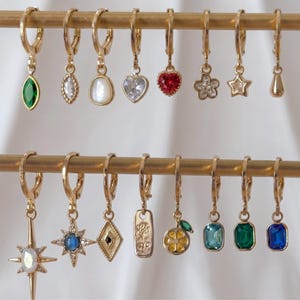 May include: A collection of sixteen gold-toned hoop earrings with various dangling charms, including faceted green, blue, and red gemstones, a white pearl-like stone, a heart, a star, a flower, a rose-engraved rectangle, and a citrus slice.