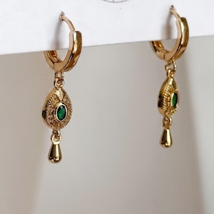 May include: A pair of gold hoop earrings featuring a dangling teardrop-shaped charm. Each charm has a textured surface with a small, oval-shaped green stone set in the center and a smooth, polished gold drop hanging at the very bottom.