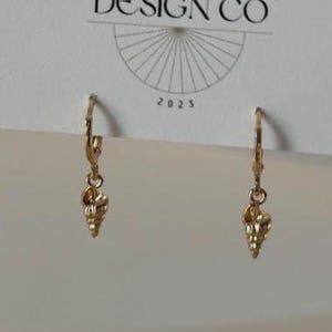 May include: Gold-colored hoop earrings with a seashell charm. The earrings are displayed on a white card with the text "JEWELRY & THINGS" and "BIG LITTLE DESIGN CO." The year "2023" is also visible on the card.