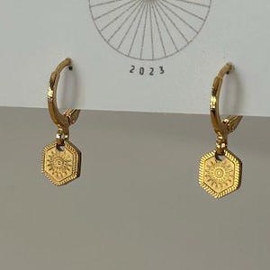 May include: Gold-colored hoop earrings with a hexagonal pendant featuring a sunburst design. The earrings are displayed on a white card with the text "BIG LITTLE DESIGN CO" and the year "2023".