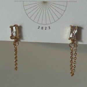 May include: Gold-toned earrings with a rectangular, clear stone and a delicate chain. The earrings are displayed on a white card with the text "BIG LITTLE DESIGN CO" and the year "2023".