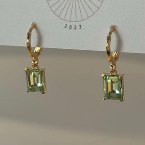May include: A pair of gold-colored hoop earrings with rectangular, light green gemstones. The earrings are hanging from a white card with the text "BIG LITTLE DESIGN CO" and the year "2023".