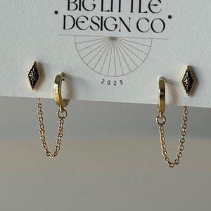 May include: Gold-colored earrings with a chain and a small black diamond-shaped stud. The earrings are displayed on a white card with the text "BIG LITTLE DESIGN CO" and "JEWELRY & THINGS". The year "2023" is also visible.