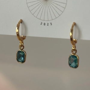 May include: A pair of gold-colored hoop earrings with a rectangular, teal-colored gemstone pendant. The earrings are displayed on a white card with the text "BIG LITTLE DESIGN CO" and "JEWELRY & THINGS" and the year "2023".