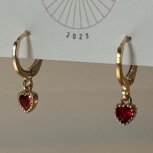 May include: Gold-colored hoop earrings with a heart-shaped, red gemstone pendant. The earrings are displayed on a white card with the text "BIG LITTLE DESIGN CO" and the year "2023". The earrings are a jewelry item.