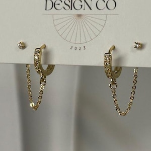 May include: Gold-toned hoop earrings with small clear stones and dangling chains. The earrings are displayed on a white card with the text "BIG LITTLE DESIGN CO" and "JEWELRY & THINGS" and the year "2023".