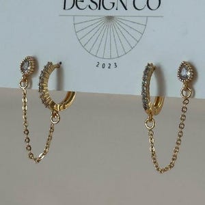 May include: A pair of gold-colored hoop earrings with small clear stones and a dangling chain. The earrings are displayed on a white card with the text "JEWELRY & THINGS" and "BIG LITTLE DESIGN CO" and the year "2023".