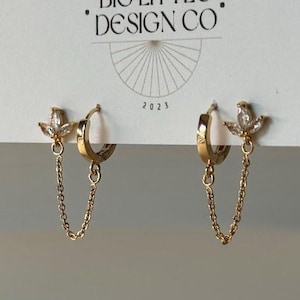 May include: A pair of gold-colored hoop earrings with a dangling chain and a floral-shaped charm with clear crystals. The earrings are displayed on a white card with the text "BIG LITTLE DESIGN CO" and the year "2023".